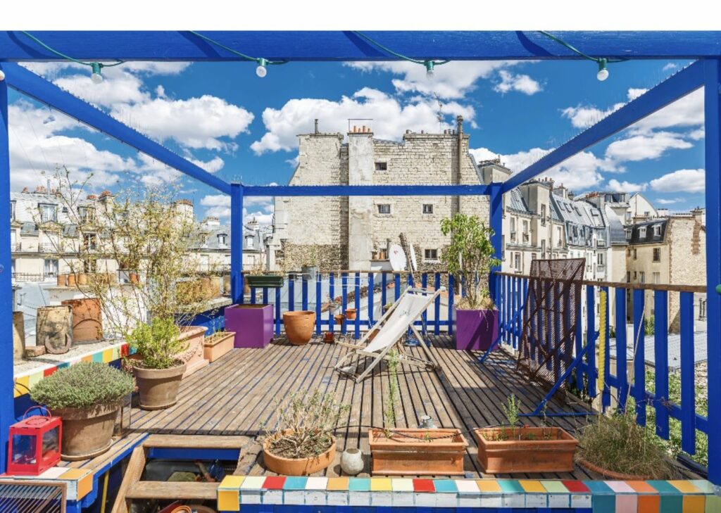 Terrasse très coloré avec une armature et une barrière bleu majorelle. Du carrelage peint de différente couleur se place autour de la terrasse. Accompagné de quelque jardinière en terre. Il y a une vue dégagé sur divers immeubles et toits de Paris. 