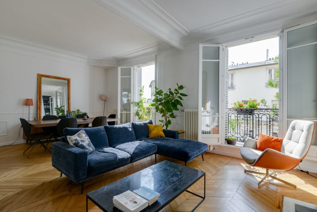 Un salon haussmannien, très chic et convivial. Appartement très lumineux, avec un canapé bleu et un fauteuil blanc et orange. 