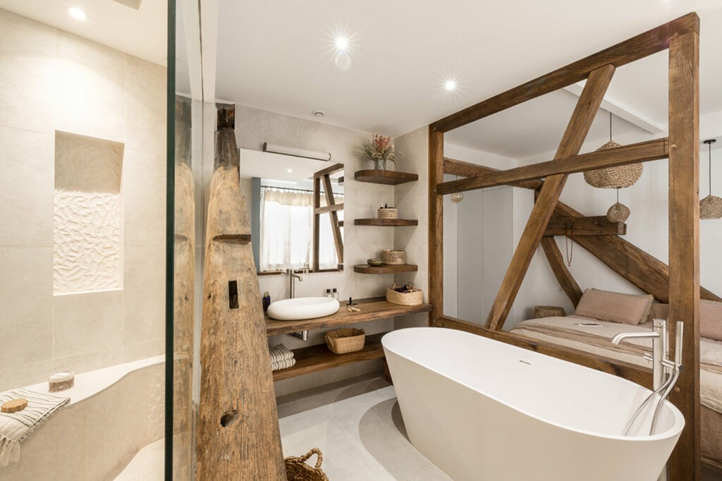 Salle de bain avec des poutres en bois disposée comme une limitation entre la chambre et la douche. Il y a une baignoire aux pieds du lit et une douche séparée d'une bais vitrée. 