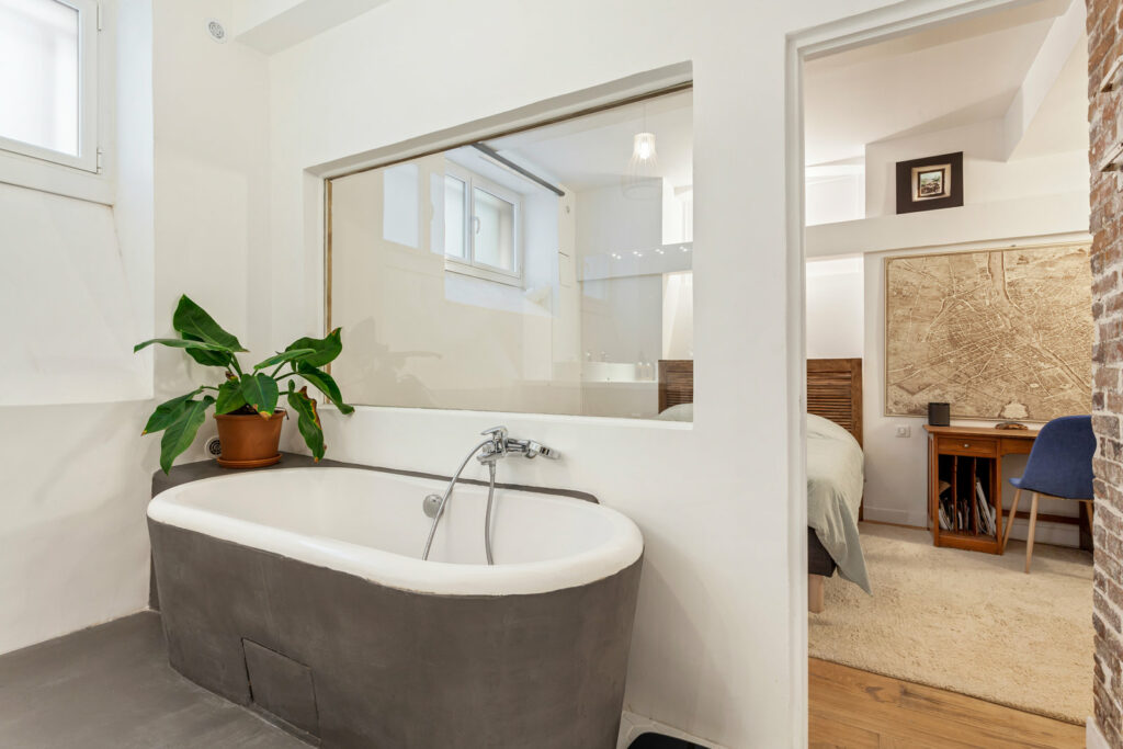 Une baignoire, couverte en extérieur de plâtre gris. Elle se situe contre un mur qui possède une vitre donnant sur la chambre. 