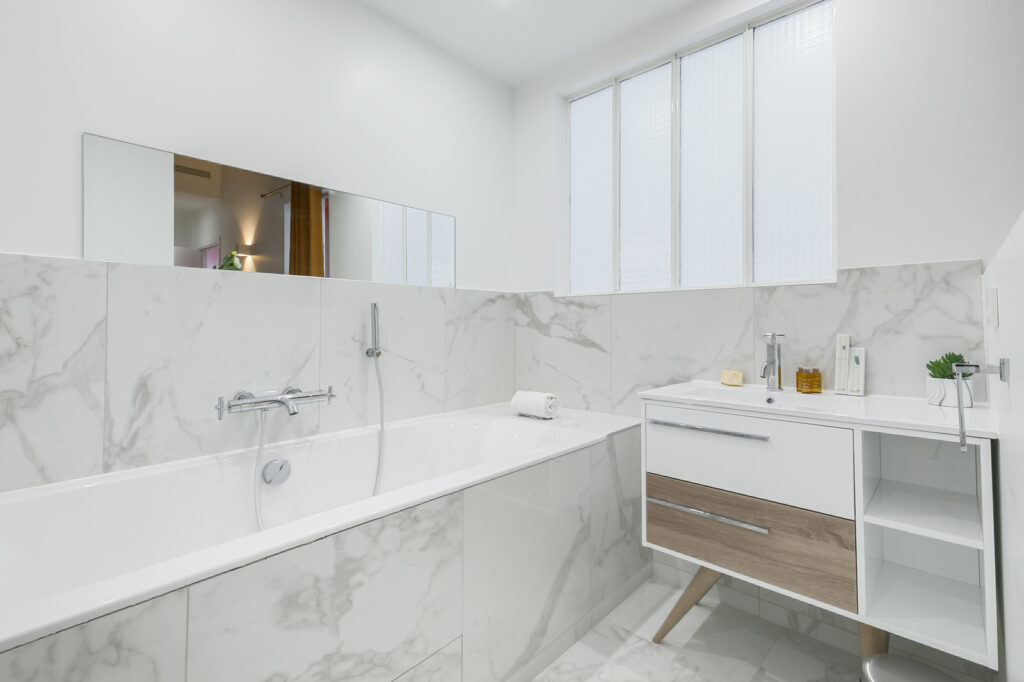 Salle de bain tout en marbre blanc. Un meuble blanc et bois avec une vasque simple est disposé à gauche de la baignoire. 