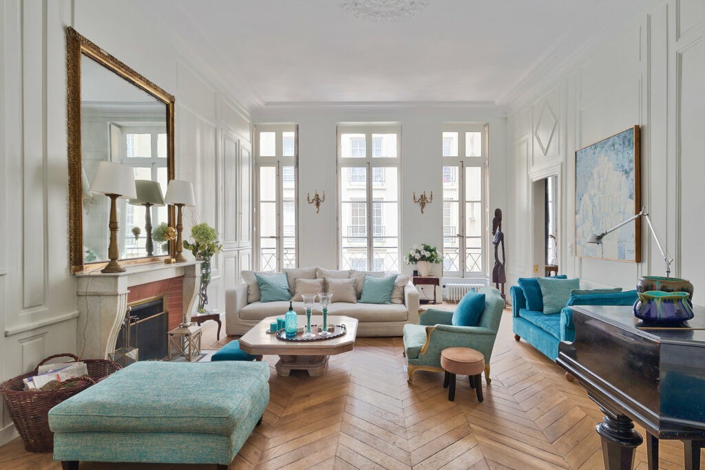Grand salon rectangulaire, peint de blanc avec du parquet au sol. il y a un grand miroir orné de dorure placé au dessus de la cheminée. les canapés sont bleue ciel placé autour d'une table basse.  